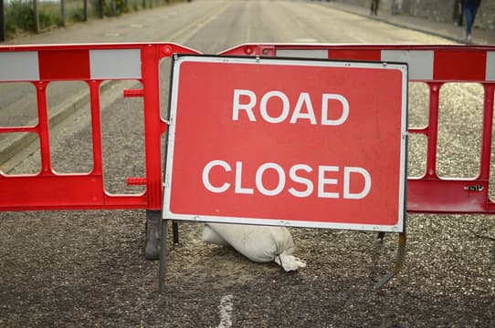 A large red sign with white letters reading ROAD CLOSED blocks a street. Red and white barriers and a sandbag support the sign, indicating that vehicles cannot pass. The road is empty in the background.