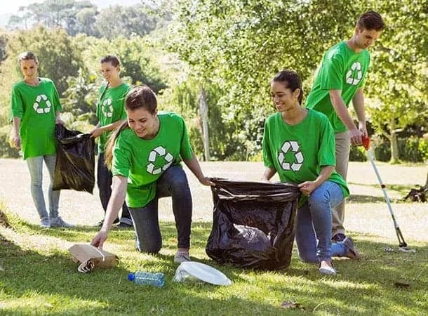 Five people wearing green shirts with recycling symbols pick up litter and place it in large rubbish bags in a sunny park with trees and grass.