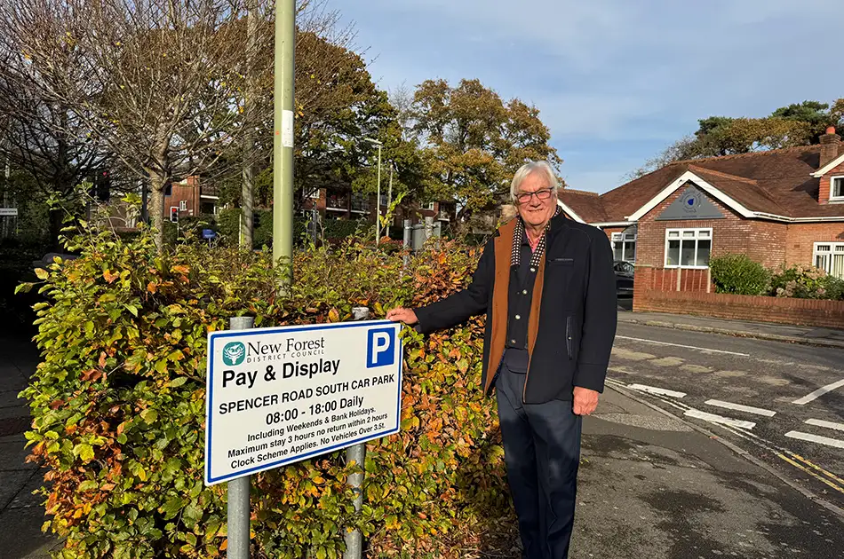 An older man with grey hair and glasses stands next to a “Pay & Display” parking sign on a sunny street corner, with houses, trees, and parked cars in the background.