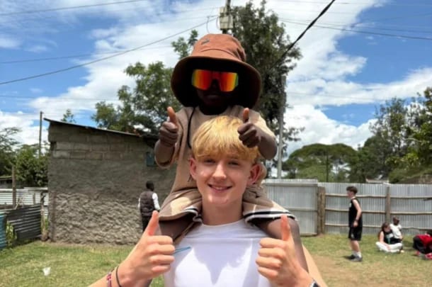 A smiling boy with blond hair gives thumbs up as a younger child wearing a brown hat, sunglasses, and matching outfit sits on his shoulders, also giving thumbs up. They’re outdoors during their life-changing trip with Lift New Forest, with people and buildings behind them.