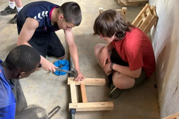 Three boys work together on a woodworking project, assembling a wooden frame on the floor—a scene inspired by student volunteering often seen in Kenyan schools. Tools and wood pieces are scattered around as they collaborate and assist each other.