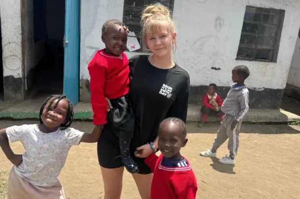 A young woman stands outside with three smiling children. Two girls wear red jumpers, and one girl wears a light-coloured dress. Other children, possibly New Forest pupils, play in the background near a weathered building with an open blue door.