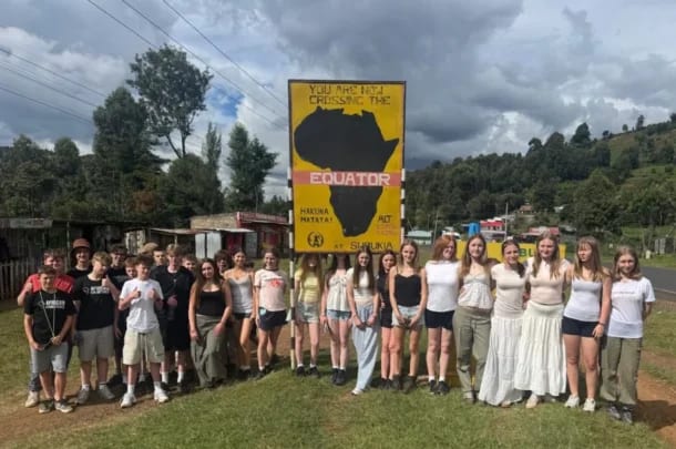 A group of young people, part of a student trip from Kenyan schools and Lift New Forest, stand smiling in front of a yellow sign that says You are now crossing the Equator with an Africa map illustration. Trees and buildings frame the cloudy sky.