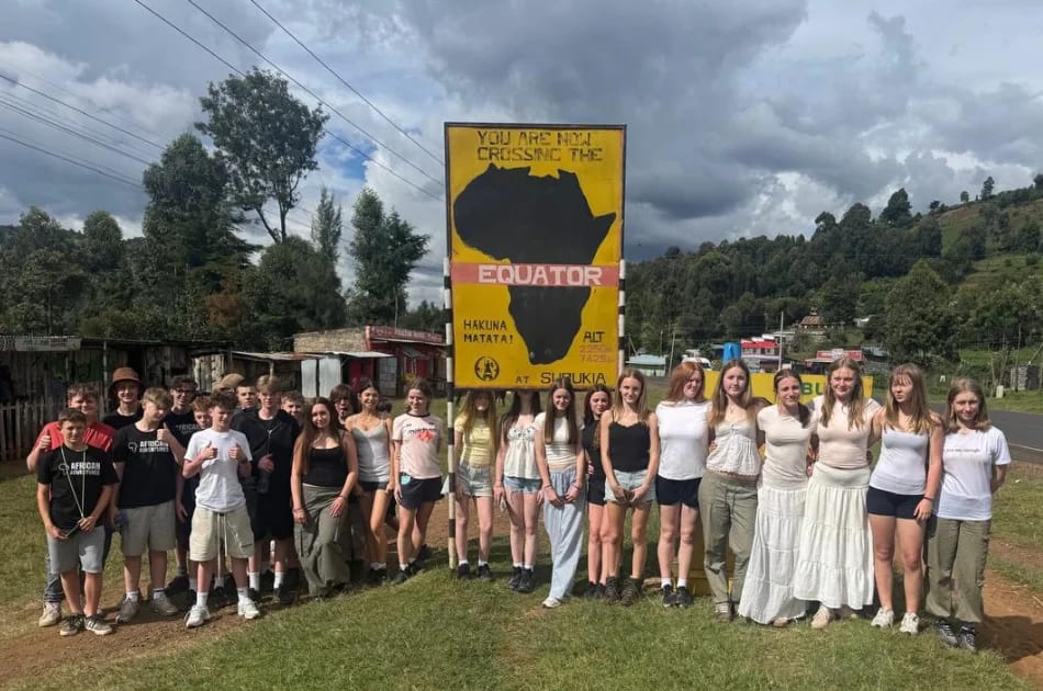 A group of young people, part of a student trip from Kenyan schools and Lift New Forest, stand smiling in front of a yellow sign that says You are now crossing the Equator with an Africa map illustration. Trees and buildings frame the cloudy sky.