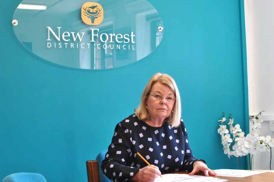 A woman with blonde hair sits at a desk, writing with a pencil, in front of a blue wall displaying the New Forest District Council sign. A vase of white flowers is on the desk beside her.