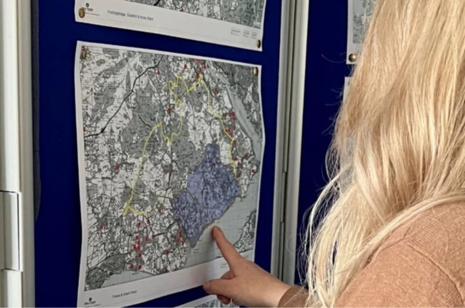 A person with long blonde hair points at a detailed map on a wall, which displays coloured lines and shaded areas, possibly for planning or analysis purposes.