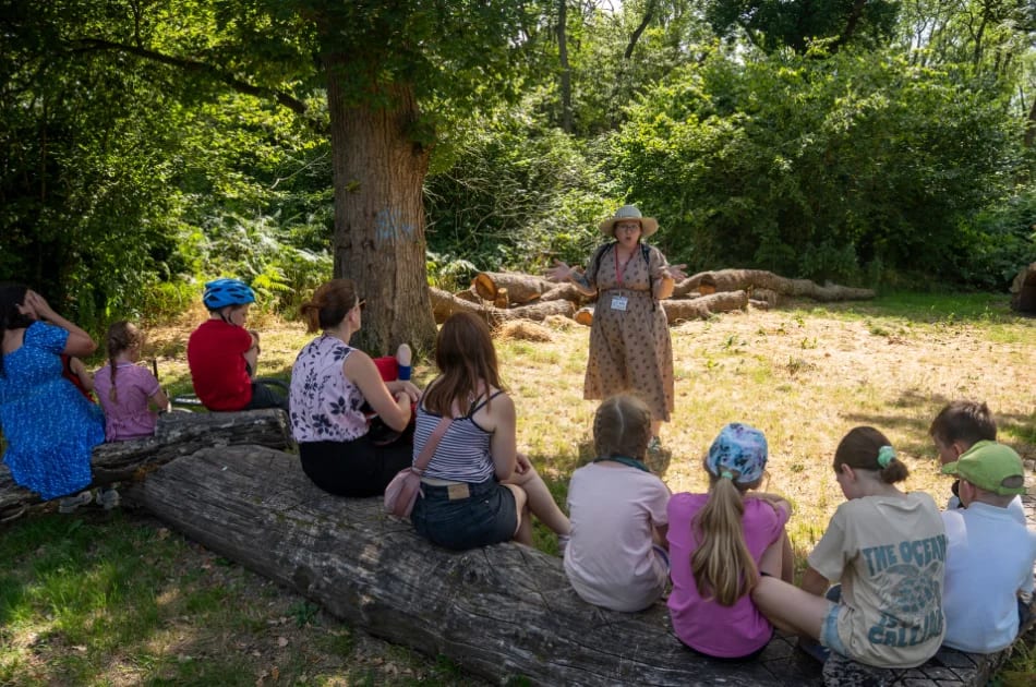 A group of children and adults sit on a large log outside, listening to a woman in a dress and hat speaking in a sunny, wooded area—a perfect setting for a Family Adventure along the enchanting Story Trails of the New Forest.