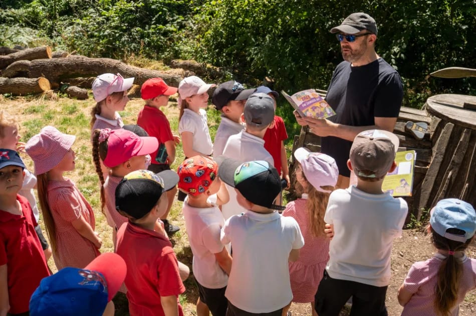 A man wearing sunglasses and a cap reads a book to a group of young children in hats outdoors. The kids, dressed in red and white, listen attentively as they enjoy this Family Adventure along Story Trails in the sunny, wooded area of the New Forest.