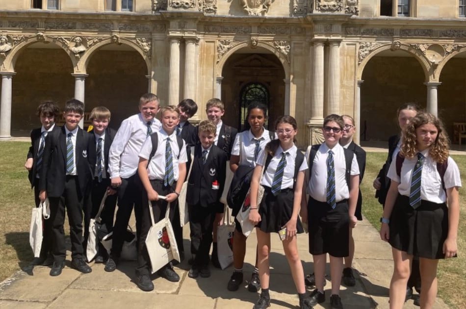 A group of school pupils in uniforms stand together outside a historic building with stone arches on a sunny day. Some carry tote bags and wear ties, while others wear skirts or shorts.