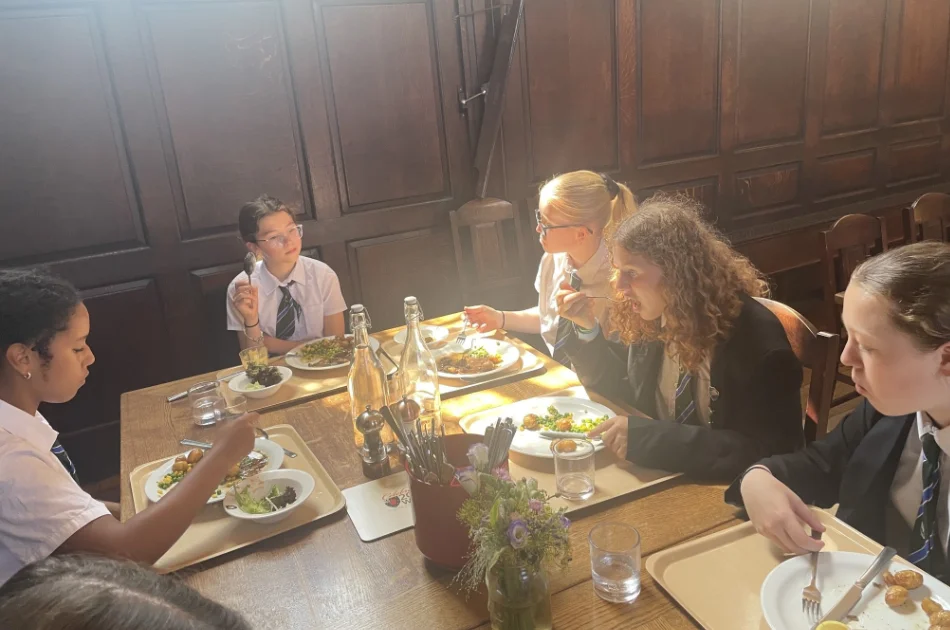 Five students in St John’s College uniforms eat lunch together at a wooden table, sunlight streaming in through a window, creating a warm, bright atmosphere. Plates of food and drinks are set on trays in front of the Year 7 friends as they chat enthusiastically about their latest exam success.