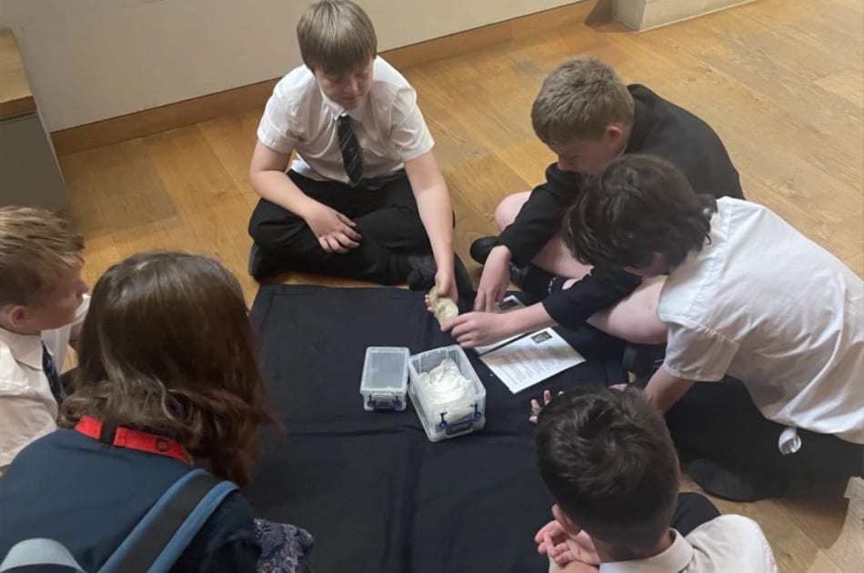 Six Year 7 pupils from St John’s College, dressed in school uniforms, sit on the floor around a black cloth, examining various objects—including a container and a rolled item. A sheet of paper with text lies on the cloth as they appear engaged in a collaborative group activity, building skills that contribute to their exam success.