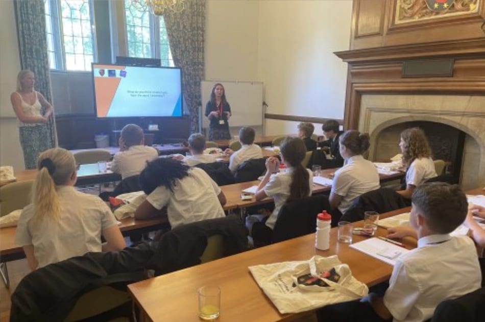 A classroom at St John’s College with Year 7 pupils in uniform sitting at tables, listening attentively to two women at the front. A screen displays a presentation on exam success, and notebooks, drinks, and bags are scattered across the tables. Large windows and a fireplace provide a welcoming backdrop.