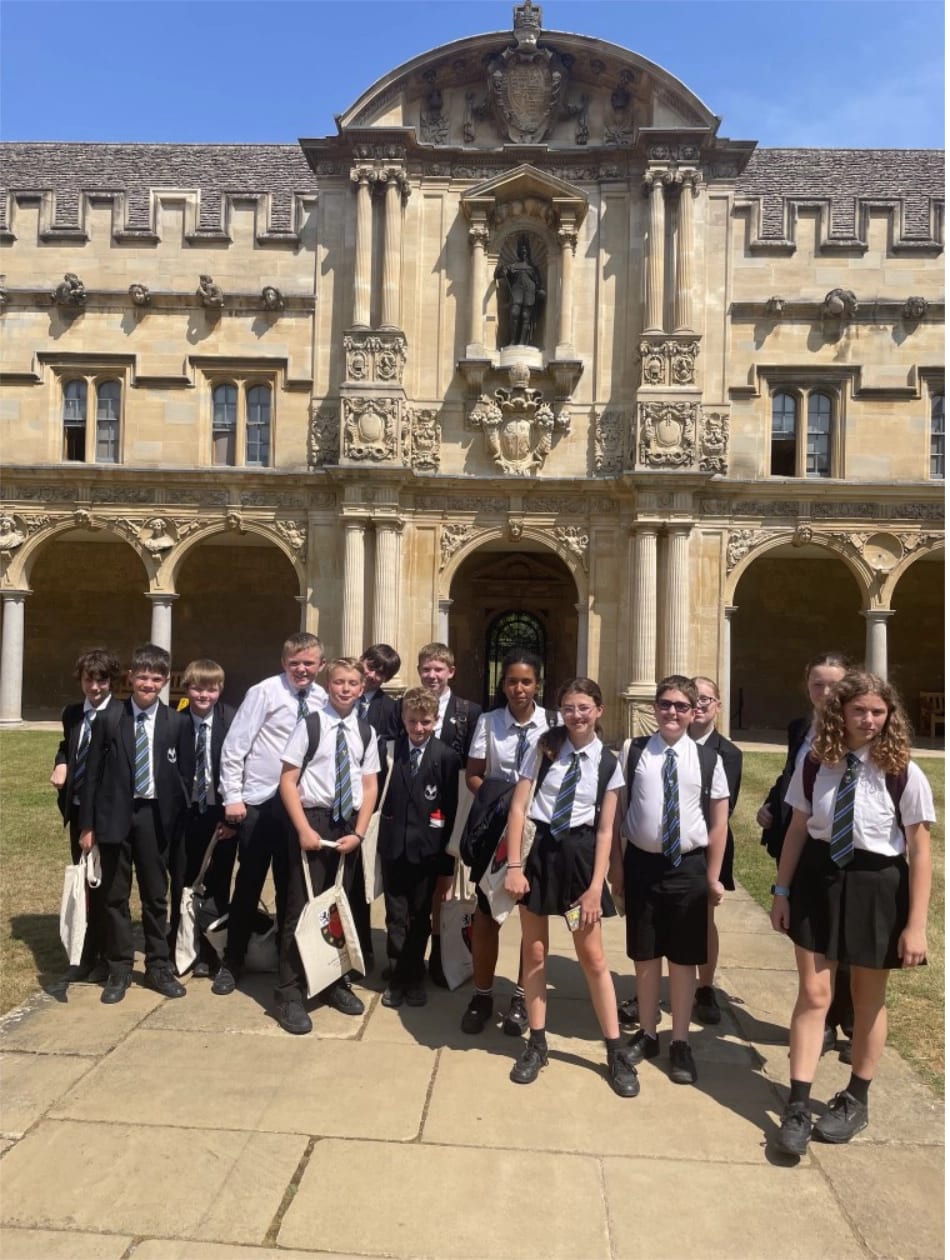A group of Year 7 pupils in school uniforms stand outside the historic stone building of St John’s College, with arched walkways and a statue above the entrance, enjoying a sunny day.