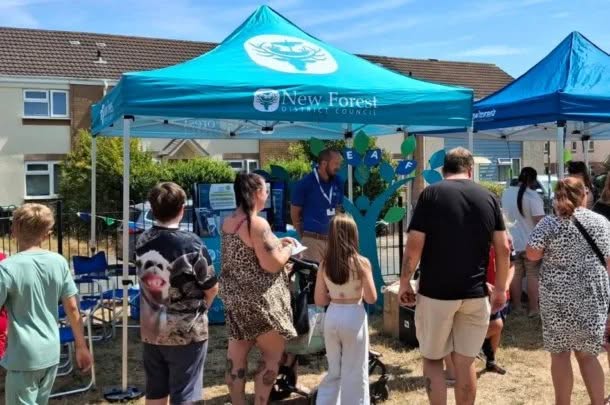 People gather under a blue New Forest District Council marquee at an outdoor event on a sunny day. Some participants wear animal print clothing and children are present. The council provides information on tenant services, and houses are visible in the background.