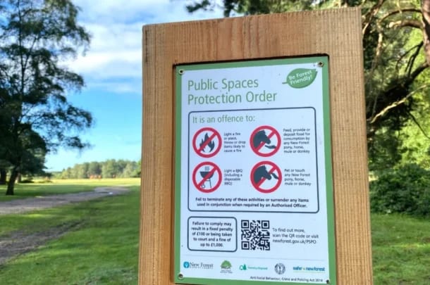 A wooden sign in a park displays rules prohibiting barbecues, fires, feeding or touching ponies and donkeys, with penalty information, set against a background of grass, trees, and partly cloudy sky.